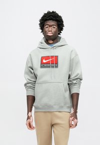 Γκρι φούτερ με κουκούλα και μπροστινή τσέπη, με ένα κόκκινο και μαύρο λογότυπο της Nike. Ριμπ μανσέτες και τελείωμα, φορεμένο με μπεζ παντελόνια.