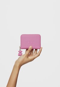 Portafoglio in pelle rosa con logo "TOUS" in rilievo, chiusura con zip e charm a forma di orsetto in silicone attaccato alla cerniera. Texture liscia, forma rettangolare.