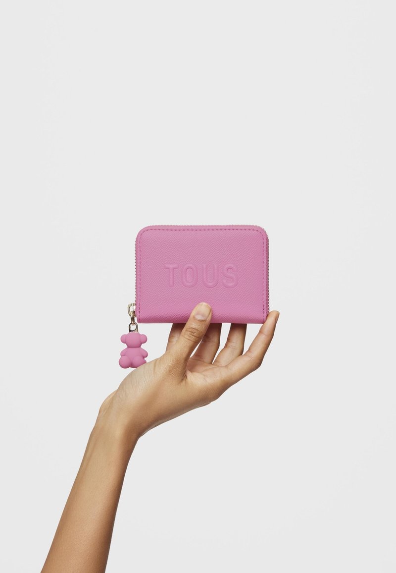 Portafoglio in pelle rosa con logo "TOUS" in rilievo, chiusura con zip e charm a forma di orsetto in silicone attaccato alla cerniera. Texture liscia, forma rettangolare.
