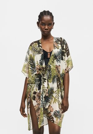 Naine punutud juustega, kellel on kuldsed rõngakujulised kõrvarõngad ja roheline, pruun ja must troopiliste lehtede mustriga kattekleit musta ujumistrikoo kohal.