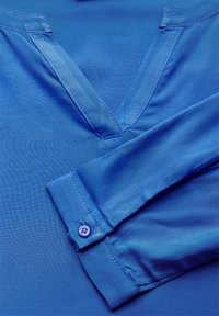 Camicia blu a V in tessuto morbido, con un colletto dettagliato e polsini abbottonati. Il materiale presenta una leggera lucentezza e texture.