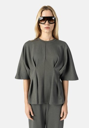 Femme aux longs cheveux blonds portant de grandes lunettes de soleil sombres et un haut gris ample à manches courtes avec un pantalon assorti, debout devant un fond blanc.
