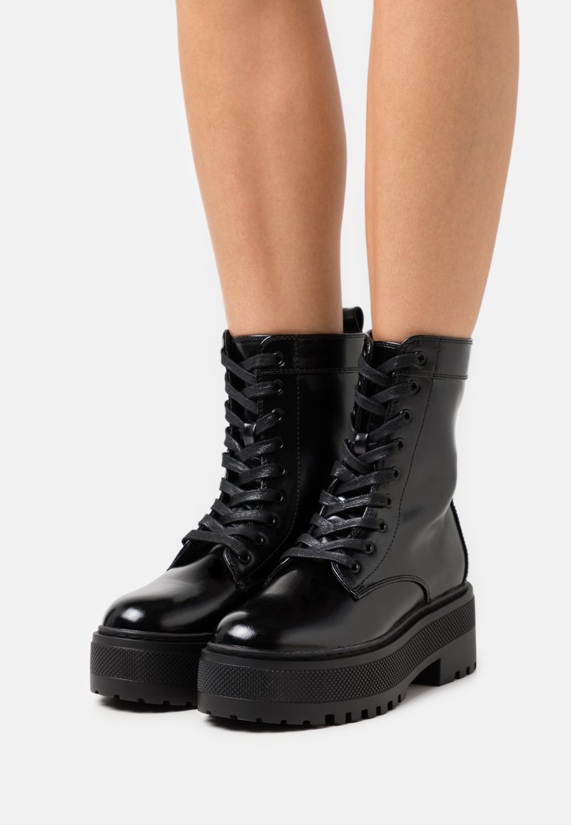 Bottes de combat plateforme noires et brillantes avec semelles épaisses et lacets, portées sur des jambes nues sur fond blanc.