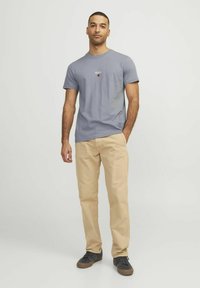 Tricou gri din bumbac cu mâneci scurte, având un mic logo "R.D.D." și un punct roșu. Asortat cu pantaloni bej și adidași negri.