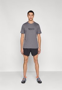 Γκρι βαμβακερό t-shirt με μαύρο τυπωμένο λογότυπο, συνδυασμένο με μαύρο σορτς και γκρι παπούτσια με πορτοκαλί λεπτομέρειες. Το μοντέλο στέκεται όρθιο.