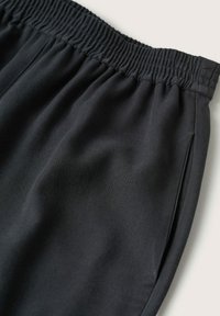 Un pantalon de survêtement noir présente une taille élastique, des poches latérales, un tissu doux et une coupe décontractée. La surface a une texture lisse.