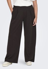 Pantalones de pierna ancha en tela negra con pliegues y bolsillos laterales. Combinados con zapatillas casuales color crema para un look relajado.
