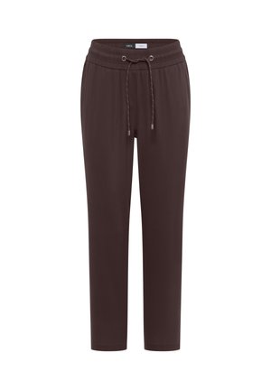 Femme souriante portant un haut bleu sans manches, un pantalon marron à cordon, tenant un grand sac tissé, debout devant un mur beige.