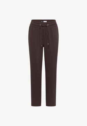 Femme souriante portant un haut bleu sans manches, un pantalon marron à cordon, tenant un grand sac tissé, debout devant un mur beige.