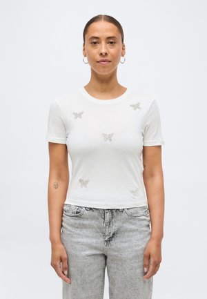 Naine, kellel on juuksed tagasi seotud, kannab valget lühikese varrukaga särki, millel on viis hõbedast liblikamotiivi, ning heledaid halli tooni pükse, seisab valge tausta ees.