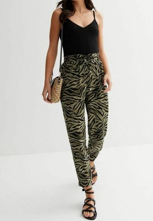 Femme portant un haut noir à fines bretelles, un pantalon vert et noir à imprimé zèbre, des sandales noires à brides et un sac bandoulière tressé.