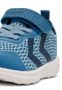 Zapatillas de malla azules con una correa de velcro, cuentan con detalles en marino y una suela blanca de goma, superficie texturizada y una marca prominente.