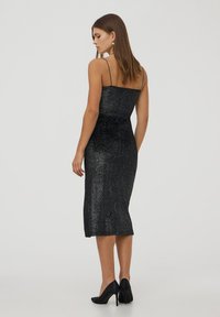 Abito midi di glitter nero con spalline sottili, forma aderente, texture lucida e una leggera svasatura all'orlo. Indossato con eleganti tacchi neri.