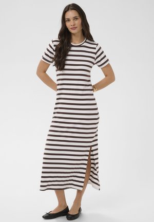 Mujer de pie con las manos detrás de la espalda, vistiendo un vestido largo de manga corta a rayas blancas y marrón oscuro con una abertura lateral y zapatos planos negros.