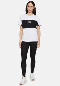 Kvinna som står i en vit och svart FILA t-shirt, svarta leggings och vita sneakers mot en enfärgad ljus bakgrund.