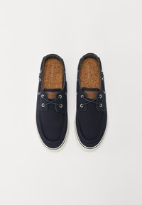 Zapatillas de lona navy sin cordones con suelas de goma blanca, que cuentan con acentos de cuero marrón y ojales de metal para los cordones. Plantillas de corcho.