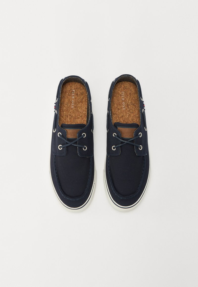 Zapatillas de lona navy sin cordones con suelas de goma blanca, que cuentan con acentos de cuero marrón y ojales de metal para los cordones. Plantillas de corcho.