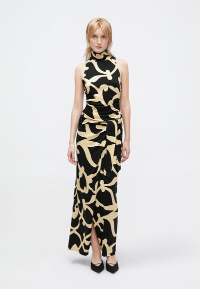 Mulher a usar um vestido maxi sem mangas preto e bege com um padrão, com um decote elevado, detalhe de fita na cintura e saltos pretos de bico fino.