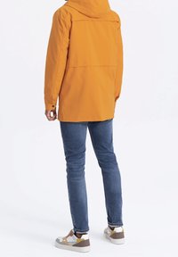 Veste imperméable jaune avec capuche, coupe décontractée et détails sur le yoke arrière, associée à un jean bleu et des baskets multicolores.