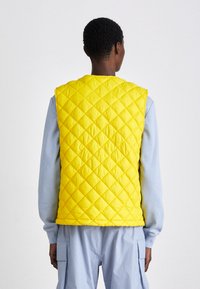 Gilet matelassé jaune avec un col haut, dotée de coutures diagonales et d'un design léger, porté par-dessus un haut à manches longues bleu clair.