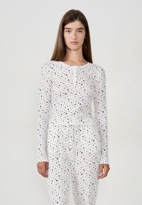 Młoda kobieta w białej, prążkowanej bluzce z długim rękawem i dopasowanych spodniach z małymi czarno-różowymi wzorami w serca, stojąca na jednolitym tle.