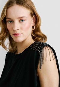 Top senza maniche nero con spalla decorata, caratterizzato da paillettes e dettagli in frange metalliche. Tessuto morbido, vestibilità comoda, design minimale.