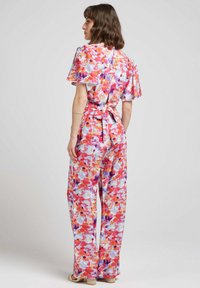 Combinaison florale avec des manches courtes en volants, une taille cintrée ornée d'un nœud, et des couleurs vives de rose, orange et violet sur un fond clair.