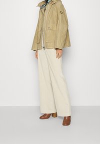 Světle hnědá voskovaná bunda s kapucí, zapínáním na zip a bočními kapsami, spárovaná se smetanově bílými kordárovými kalhotami s širokými nohavicemi a hnědými kotníkovými botami na podpatku.