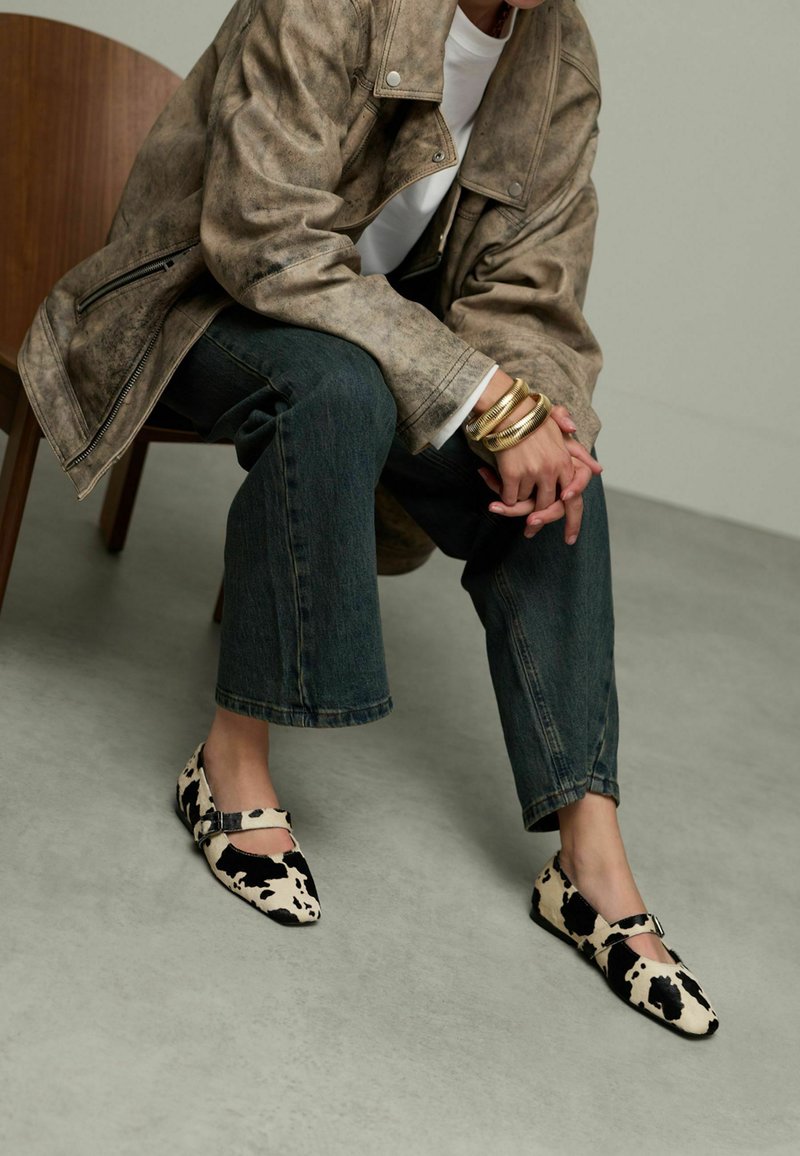 Chaussures plates à imprimé vache avec un bout pointu et une bride à la cheville, fabriquées en tissu doux. Portées avec un denim foncé, offrant une coupe décontractée et courte.