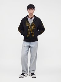 Young man wearing a black hoodie with a yellow astronaut design, light blue jeans, black and white sneakers, standing with hands in pockets.