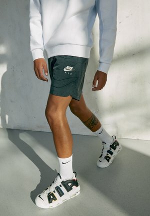 Osoba nosząca biały sweter, czarne spodenki Nike Air, białe skarpetki Nike oraz białe buty Nike Air, chodząca wewnątrz na szarej podłodze.