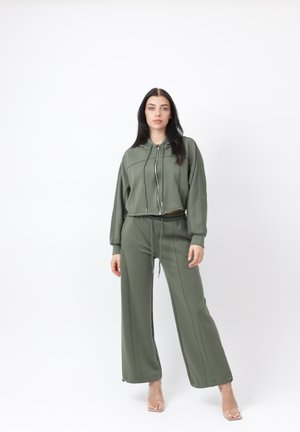 Vrouw staand, gekleed in een olijfgroene zip-up hoodie met trekkoorden en bijpassende wijde broek met een trekkoord in de taille, transparante hoge hakken.