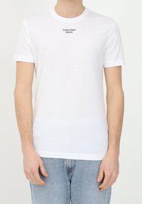 Bílá bavlněná tričko s krátkými rukávy, kulatým výstřihem a černým logem "Calvin Klein Jeans" umíst�ěným uprostřed na prsou. Hladká textura.