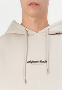 Beige Kapuzenpullover aus weichem Stoff mit einer Kängurutasche, verstellbarem Kapuzenzug und einem schwarzen Logoaufdruck auf der Vorderseite.