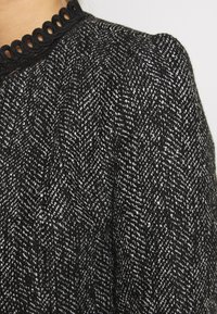 Sort og hvid fiskebenmønsterstof med en tekstureret finish og en sort bølget kant langs halsudskæringen.