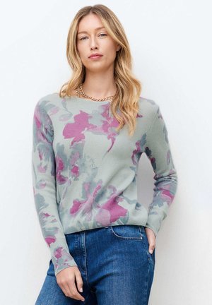 Kobieta o długich blond włosach, ubrana w szary i różowy sweter w kwiaty, niebieskie dżinsy oraz złoty łańcuszek, pozująca na białym tle.