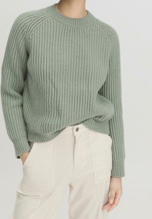 Femme portant un pull côtelé vert clair rentré dans un pantalon taille haute écru, les mains dans les poches, debout devant un fond uni.