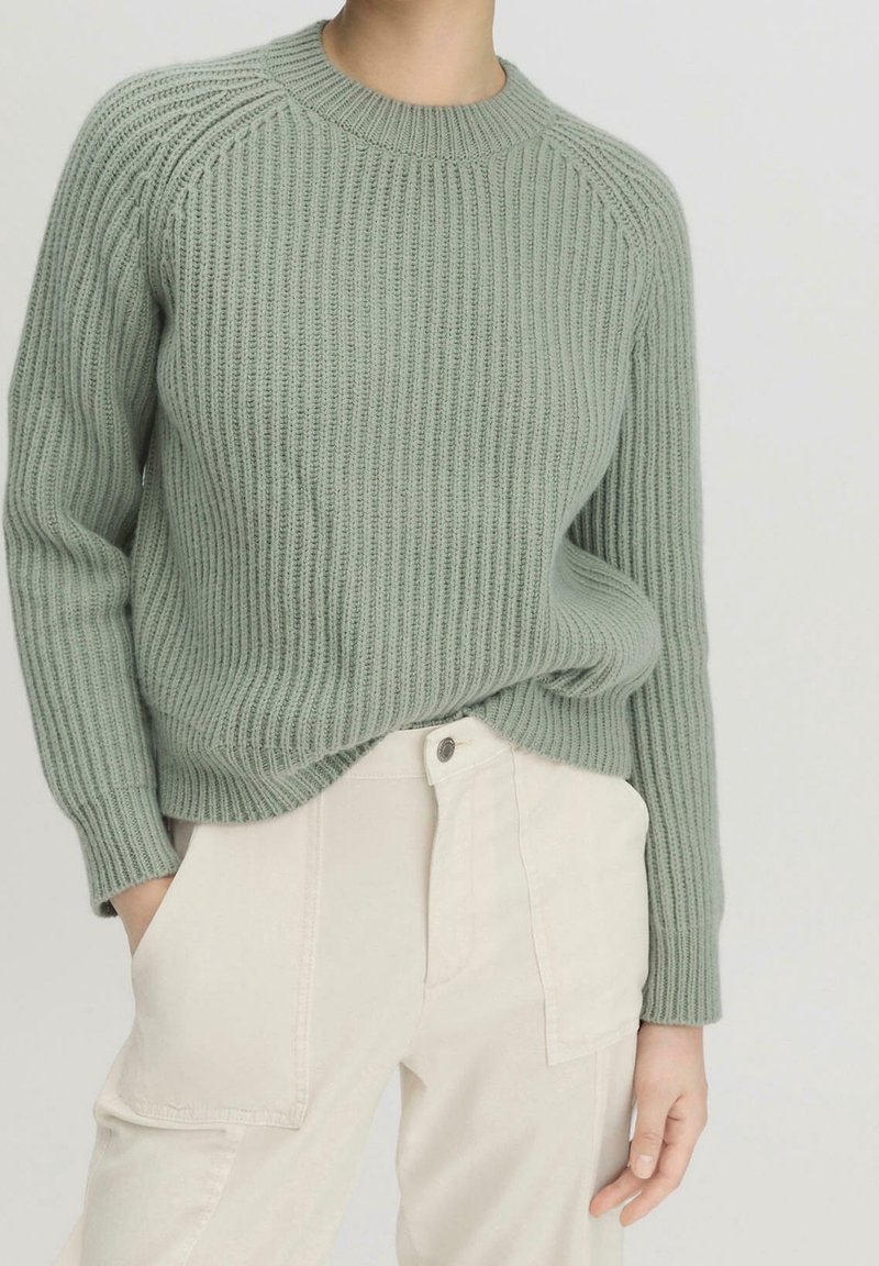 Femme portant un pull côtelé vert clair rentré dans un pantalon taille haute écru, les mains dans les poches, debout devant un fond uni.