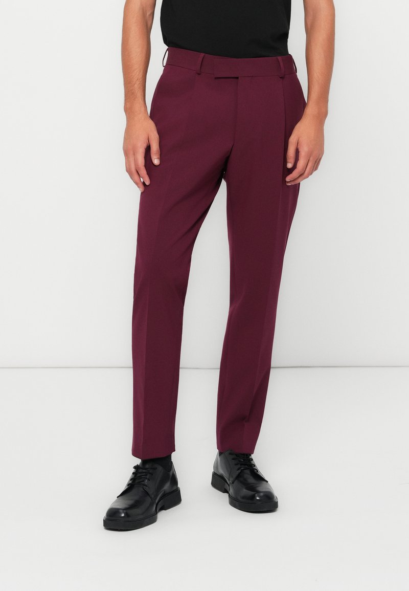 Pantaloni su misura bordeaux realizzati in tessuto liscio, con frontale piatto, tasche laterali e vestibilità slim, abbinati a scarpe nere.