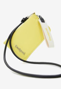 Geel synthetisch schoudertasje met een driehoekige vorm. Voorzien van een zwarte leren band en zilveren hardware. "Desigual" logo in reliëf op de voorkant.