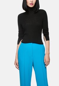 Frau, die einen schwarzen Rollkragenpullover mit Dreiviertelärmeln und leuchtend blauen, hochgeschnittenen Hosen vor einem weißen Hintergrund trägt.