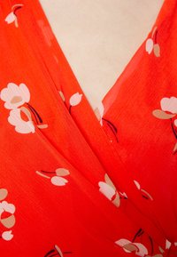 Tissu rouge avec un motif floral dans des tons de rose pâle et beige. Design drapé avec un col en V, présentant une apparence légère et texturée.