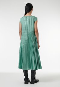 Faltendes grünes Satin-Kleid mit kurzen Ärmeln, Detail mit Schlüsselöffnung im Rücken und luftiger Silhouette, kombiniert mit schwarzen Ankle Boots.