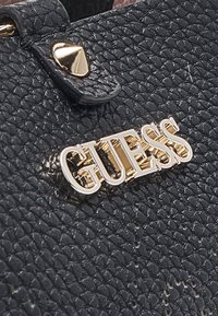 Matériau en cuir noir texturé avec un logo "GUESS" signature en or, comprenant un accent métallique et une petite rivet décorative.