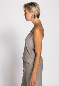 Woman with short blonde hair in taupe sleeveless top and pants, standing in profile against a plain light background.