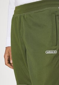 Närbild av gröna Adidas-byxor med elastiskt midjeband och sidficka, som bärs tillsammans med en vit långärmad tröja.