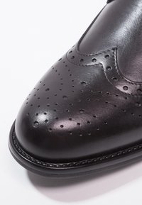 Chaussure brogue en cuir noir avec détails perforés, design courbé, texture brillante et bordure cousue le long de la semelle.