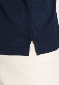 Granatowy sweter dzianinowy z teksturowanym wykończeniem, z tylnym rozporkiem i prążkowanym wykończeniem dołu. Ułożony na jasnym tle.