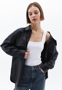 Zwarte faux leren shirt met een knoopsluiting, grote klepzak en lange mouwen, gedragen over een witte tanktop en blauwe jeans.