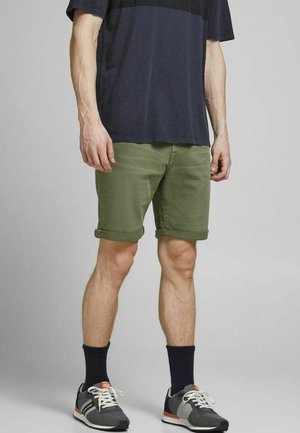 Pantaloni corti verdi in cotone con orli risvoltati, indossati con una camicia blu navy e sneaker grigie. Caratterizzati da calzini scuri e un design semplice.